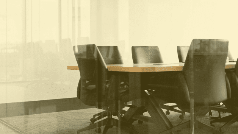 Empty conference room with black office chairs around a wooden meeting table seen through a glass wall.