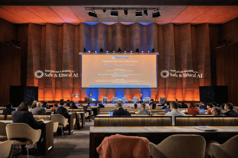 Auditorium with attendees facing a large stage and presentation screen during a conference on safe and ethical AI.
